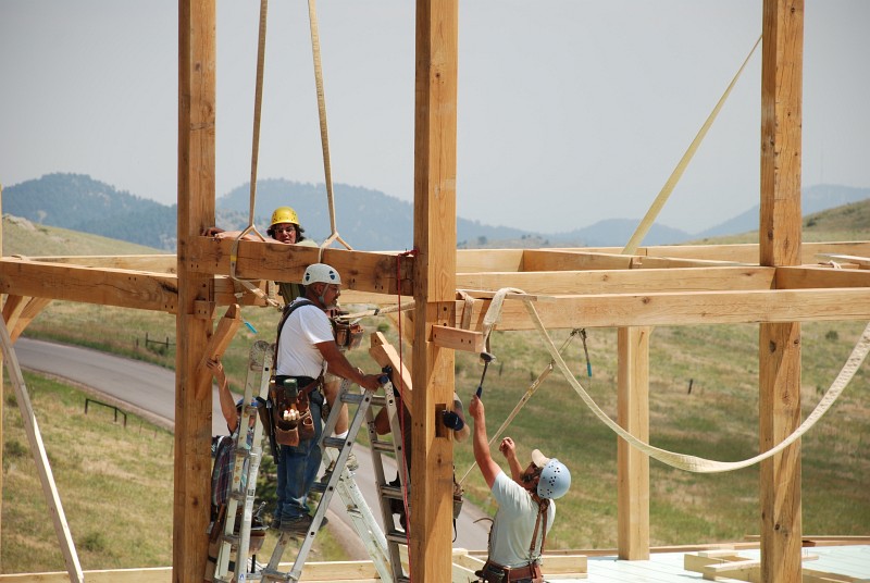 A beam is being fitted between bents 6 and 5. It takes a lot of help to get the beam in place: Matt is guiding the east end onto the floating tenon, below him Jason is holding the brace in place, Jaime (white helmet) is guiding the west end onto the other floating tenon, Harris (behind the post) is holding the other brace in place, and Darby is pulling a lift strap out of the way. It takes a lot of people to set just one beam!